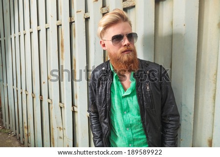young casual redhead bearded man looking up, away from the camera, while posing outdoor, by a rusty iron fence