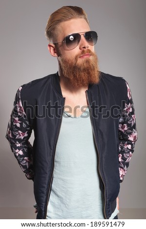 casual young man with a long red beard holding both hands in his rear pockets and looking away from the camera. on gray studio background