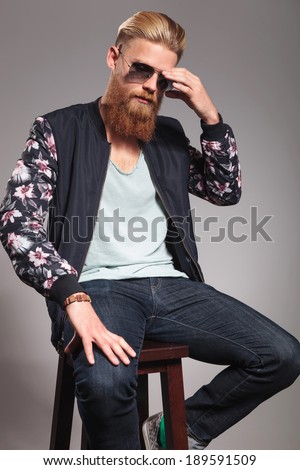 casual young man with a long red beard sitting on a high stool and adjusting his sunglasses while looking down, away from the camera. in a gray background studio