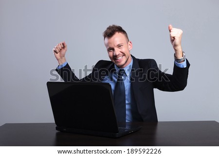 young business man cheering in front of the laptop while looking into the camera. on a gray studio background