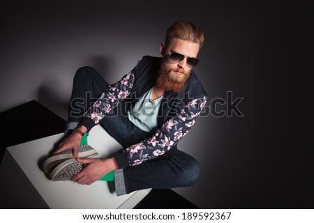 seated young bearded man looking away from the camera and holding the soles of his feet together. on a black studio background