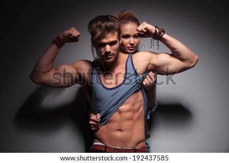 sexy woman pulls up her boyfriend\'s shirt to reveal his abdominal muscles while he is flexing his bicecps