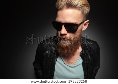 casual young man with an unshaved beard looks away while holding his hands at his back. on a dark studio background