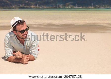 smiling young man with sunglasses and hat , lying on the beach and looks away