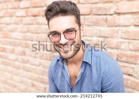 closeup picture of a happy smiling young casual man with glasses leaning against brick wall
