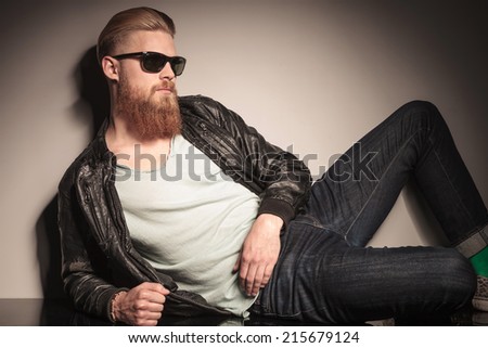 Casual young man in leather jacket pulling his sunglasses, sitting on the floor, looking at the camera