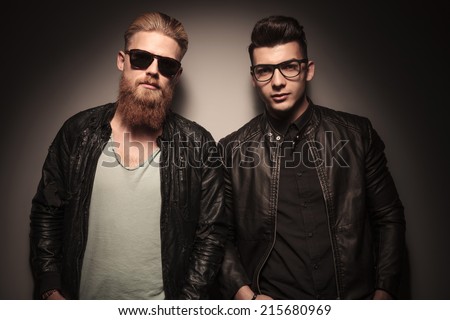 Two hot guys in leather jacket with glasses, looking at the camera, against studio background