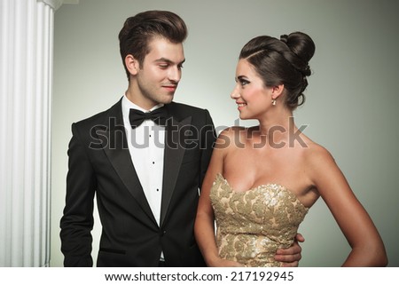 man in tuxedo embracing his woman and smiles at her near column in studio