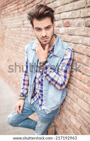 Young casual man leaning on brick wall and holding his chin with his hand, looking at the camera.