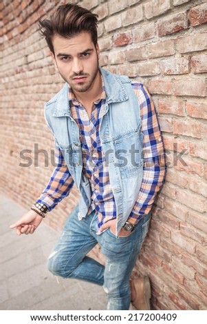 Picture of a casual handsome man leaning on a brick wall with one hand in the pocket, looking at the camera.