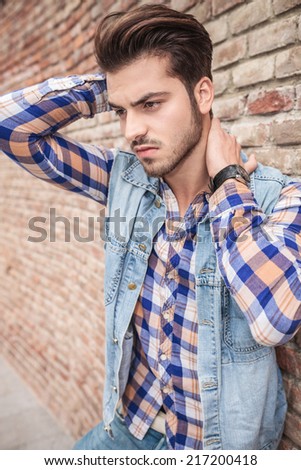 Side view of a young man leaning on a brick wall holding his hand on his neck, looking away from the camera