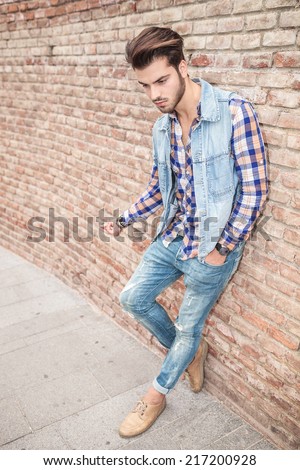 Cool fashion man in blue shirt and sunglasses sitting with his hand in pocket, smiling and looking away.