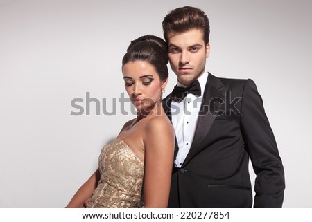 Close up of an elegant couple posing on grey studio background, the woman is leaning on her lover looking down, while he is looking at the camera.