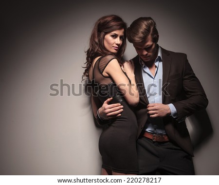 Side view of a elegant woman wearing a black dress embracing her boyfriend while he is leaning on the wall holding his jacket with one hand.