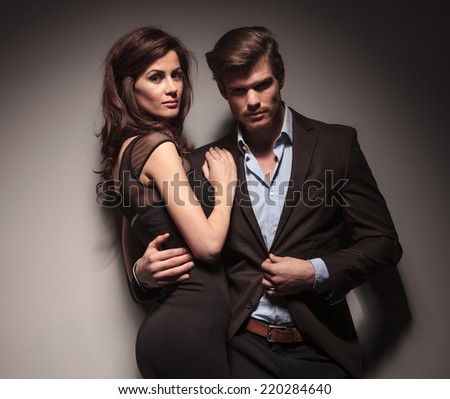 Elegant couple embracing and leaning on a dark grey wall, looking into the camera.