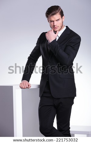 Handsome young business man leaning on a white table with one hand and scratching this beard with the other hand.