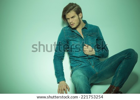 Picture of a handsome fashion man looking away from the camera while siiting on the floor, pulling his shirt.