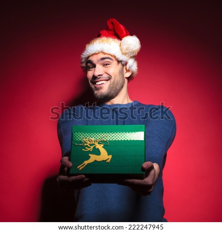 Handsome young santa giving you a green gift while smiling. On red backgrpund.