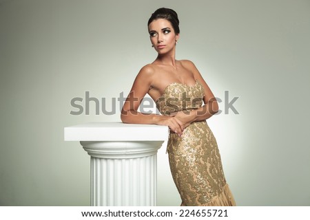 classy young woman in long elegant dress, standing near column and looks away in studio