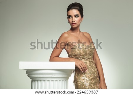 side view of a young beautiful woman in golden dress leaning on a column and looking to her back in studio