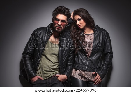 Fashion man leaning on a grey wall with his hands in pocket while his girlfriend is leaning on him.