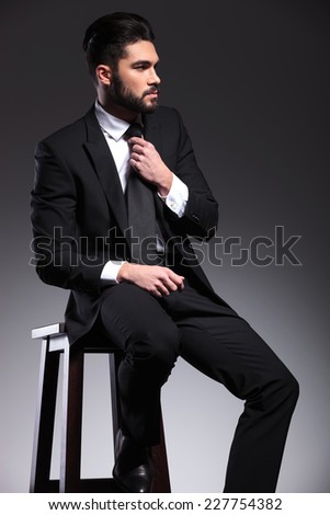 Side view of a elegant business man sitting on a stool while fixing his tie, looking away from the camera.