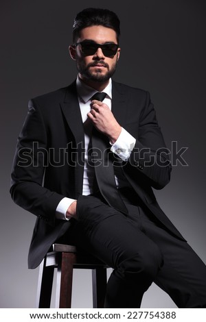 Young business man fixing his tie while holding one hand in his pocket, sitting on a stool.