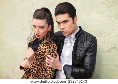 Young fashion man looking at the camera while holding his lover by the shoulder, against an old green wall.