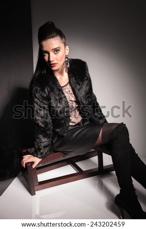 Young beautiful fashion woman leaning on a stool while sitting on it, looking at the camera.
