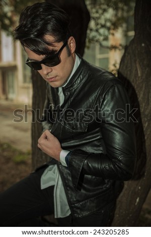 Portrait of a young casual man looking down while leaning on a tree, pulling his jacket.