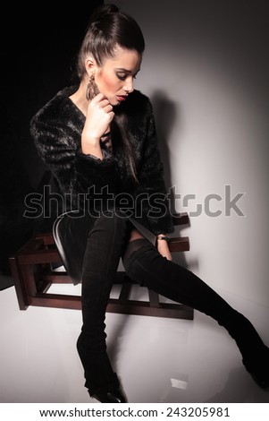 Attractive young elegant woman sitting on a stool while looking down, holding her hand to her chin.