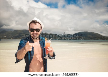 Attractive young happy man pointing at you while holding a cocktail in his hand, on the beach.