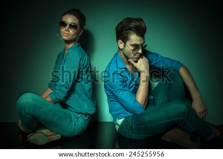 Man and fashion woman sitting back to back on studio background. she is looking at the camera while he is looking down thinking.