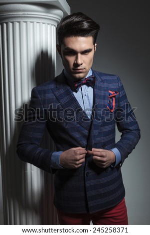 Smart casual fashion man looking down while closing his jacket, near a white column on studio background.