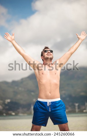 Portrait of a young happy man enjoying his vacation at the seaside, holding both hands in the air, laughing.