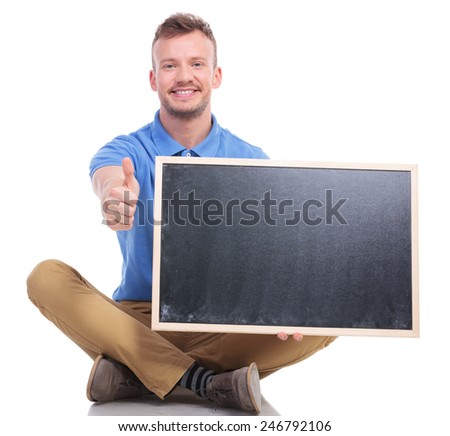 picture of a casual young man sitting on the floor with his feet crossed while holding a small blackboard and showing the thumb up gesture. on a white background