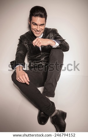 Happy young fashion man relaxing on white studio background, holding his hands on his kness while lookig at the camera.