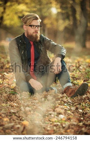 Picture of a handsome casual man sitting in the park, resting his hand on his knee while looking away from the camera.
