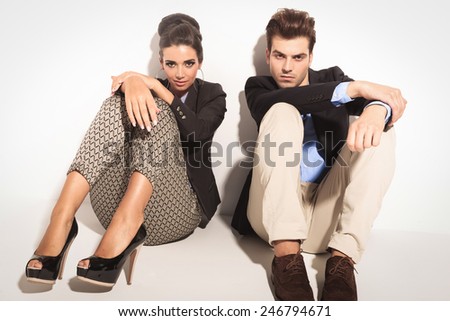 Fashion couple sitting on studio background with the knees up, holding their hands on them.