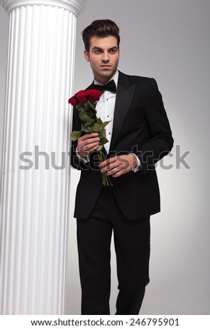 Handsome young business man posing near a white column with a bouquet of red roses in his hand, looking away from the camera.