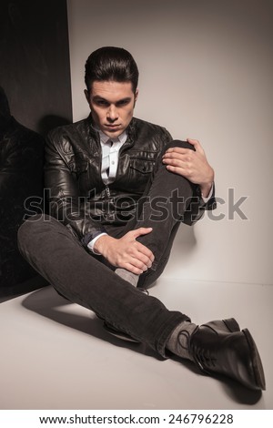 Handsome young casual man sitting on studio background holding his knee with his left hand, looking at the camera