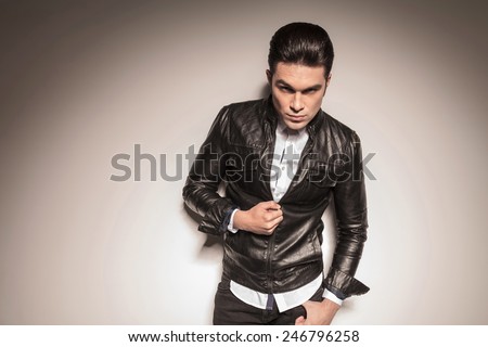 Picture of a casual young man closing his leather jacket while looking at the camera, holding his thumb in his pocket.
