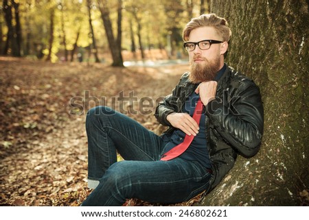Side view picture of a young casual man sitting near a tree while fixing his tie.