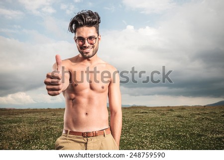 Shirtless man posing outside with his hand in his pocket while showing the thumbs up gesture.