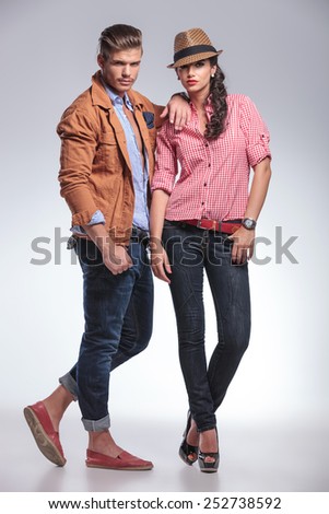 Full body picture of a young fashion couple posing on grey studio background, the man is leaning on the woman.
