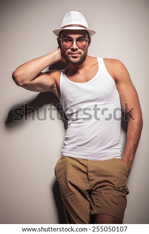 Handsome young casual man holding one hand behind his neck while leaning on a white wall with his hand in pocket.