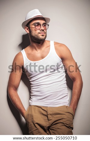 Handsome young fashion man holding both hands in his pocket while leaning on a white wall, looking away from the camera.