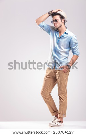 Side view picture of a young casual man posing on studio background with one hand in his pocket, fixing his hat.