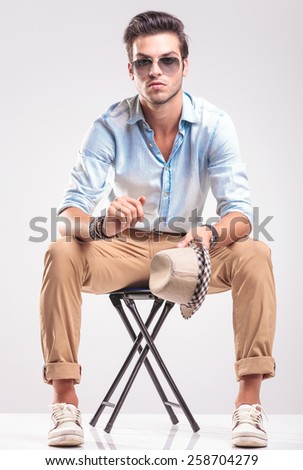 Casual fashion man sitting on a stool, holding his hat in his left hand while looking at the camera.