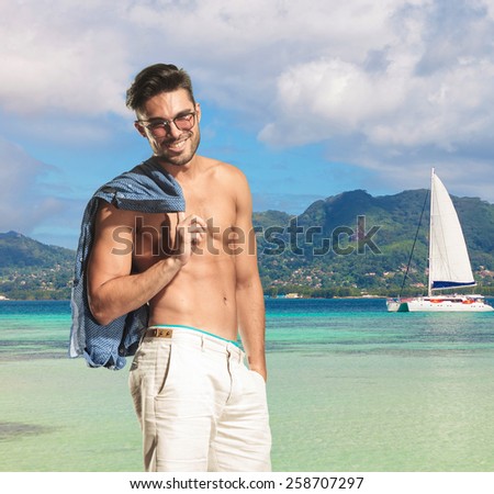 man relaxes on the shore of blue sea, holding shirt on his shoulder and smile